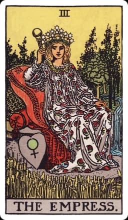 The Empress from the Rider-Waite-Smith deck: a pregnant-looking woman in a flowing gown patterned with pomegranates reclines on a cushioned outdoor throne set in a lush wheat field, a crown of twelve stars on her head, a heart-shaped shield bearing the astrological symbol for Venus resting beside her, and a stream of water flowing through trees in the background.