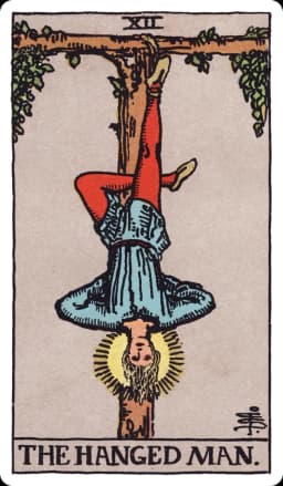 The Hanged Man from the Rider-Waite-Smith deck: a young man hangs upside down by one foot from a T-shaped wooden gibbet formed from a living tree, his other leg bent to form a numeral four, his hands folded calmly behind his back, a soft golden halo glowing around his head, his expression peaceful rather than distressed.