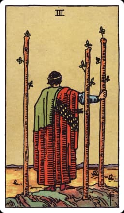 The Three of Wands from the Rider-Waite-Smith deck: a robed figure stands on a cliff edge with his back to the viewer, watching ships sail across the sea below, three tall wands planted firmly in the ground around him, one gripped lightly under his hand.
