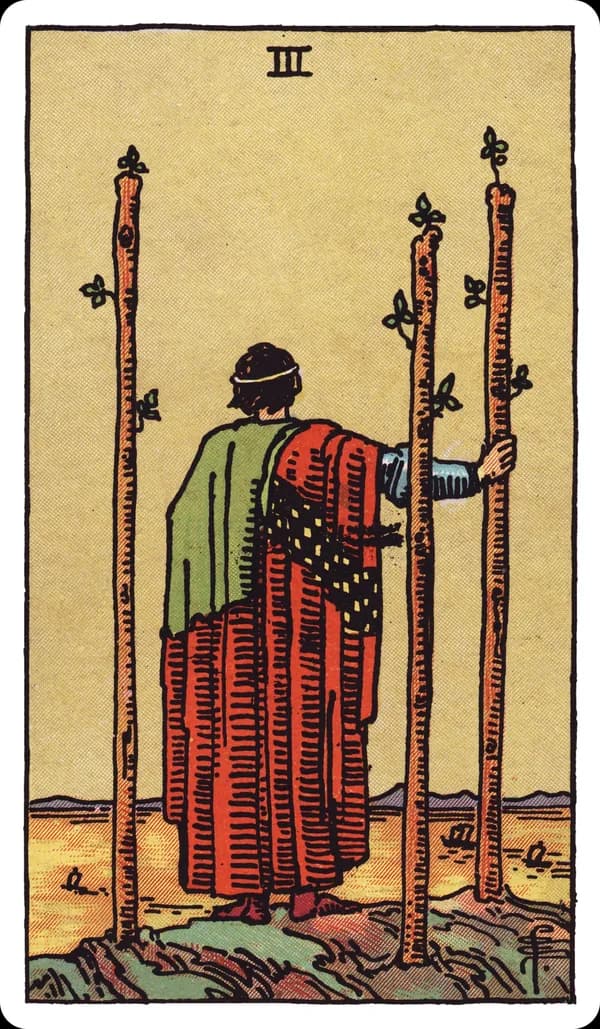 The Three of Wands from the Rider-Waite-Smith deck: a robed figure stands on a cliff edge with his back to the viewer, watching ships sail across the sea below, three tall wands planted firmly in the ground around him, one gripped lightly under his hand.