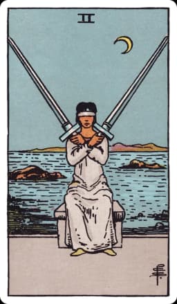 The Two of Swords from the Rider-Waite-Smith deck: a woman in pale robes sits blindfolded at the edge of a rocky shoreline, two full-length swords crossed and balanced against her shoulders, her arms folded over her chest to hold them steady, a choppy sea and jagged rocks behind her under a low crescent moon.