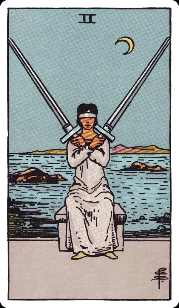 The Two of Swords from the Rider-Waite-Smith deck: a woman in pale robes sits blindfolded at the edge of a rocky shoreline, two full-length swords crossed and balanced against her shoulders, her arms folded over her chest to hold them steady, a choppy sea and jagged rocks behind her under a low crescent moon.