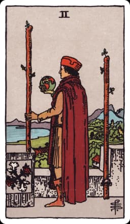 The Two of Wands from the Rider-Waite-Smith deck: a man in rich robes stands on a castle battlement overlooking sea and distant mountains, one wand fixed to the wall beside him and the other held loosely in his hand, a small globe resting in his other palm.